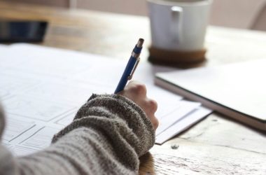 person writing on brown wooden table near white ceramic mug