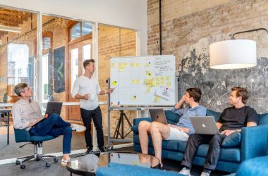 three men sitting while using laptops and watching man beside whiteboard