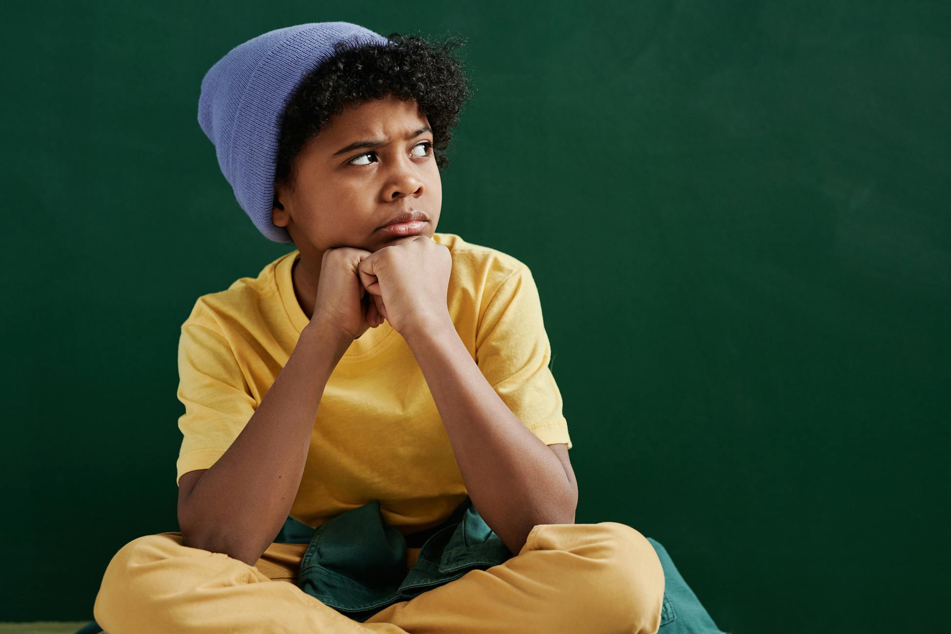 Pensive boy with curly hair wearing a yellow shirt and blue beanie against a green background.