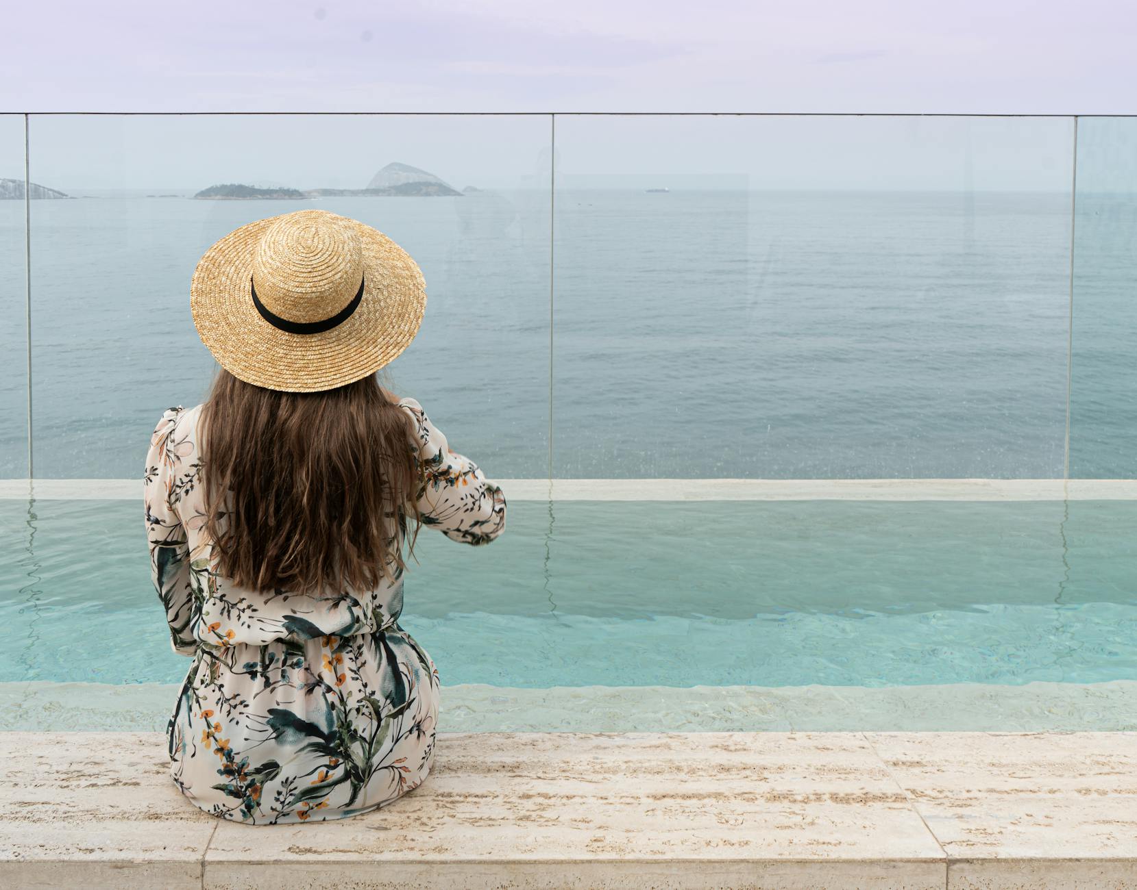 A woman in a floral dress and straw hat enjoys ocean views by an infinity pool.