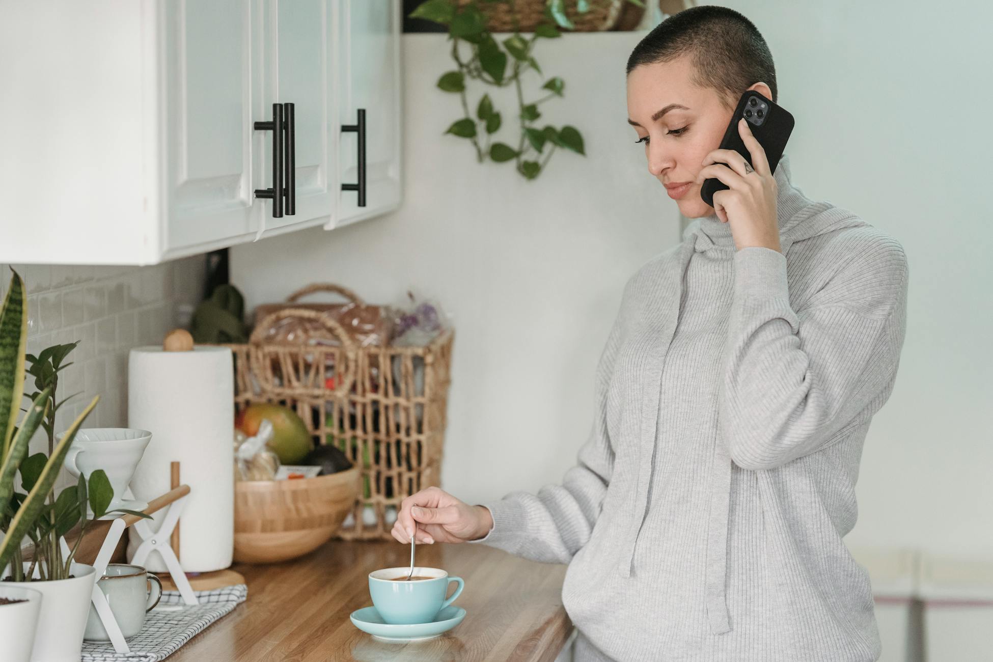 Calm young female in domestic wear having conversation on mobile phone and stirring coffee while standing in contemporary light kitchen