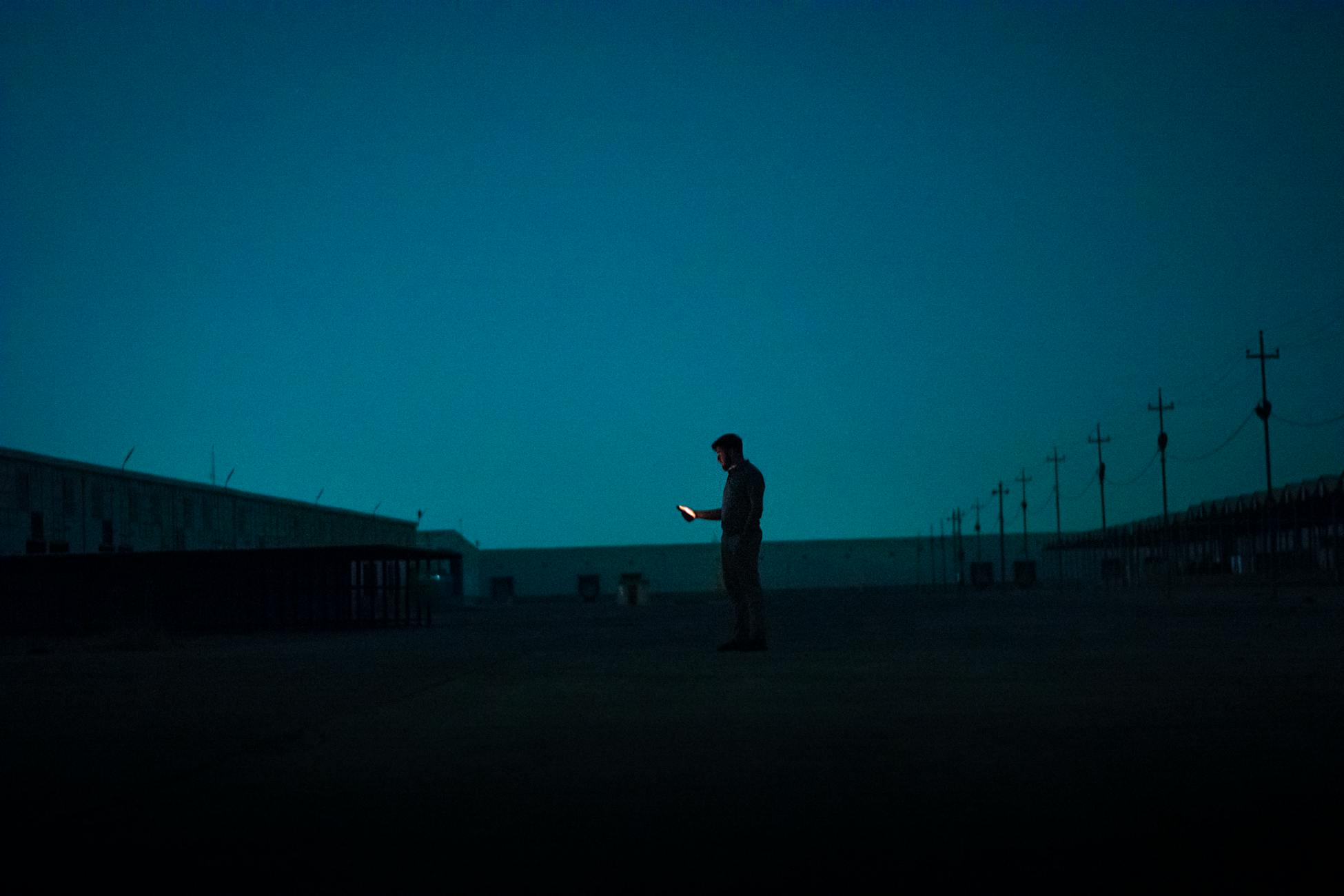 A solitary figure illuminated by a phone screen, standing alone in a dark, empty outdoor space at night.