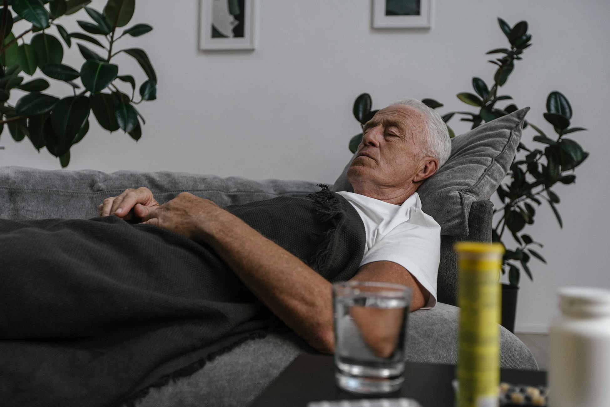 An elderly man sleeping on a couch with a blanket and glass of water nearby.
