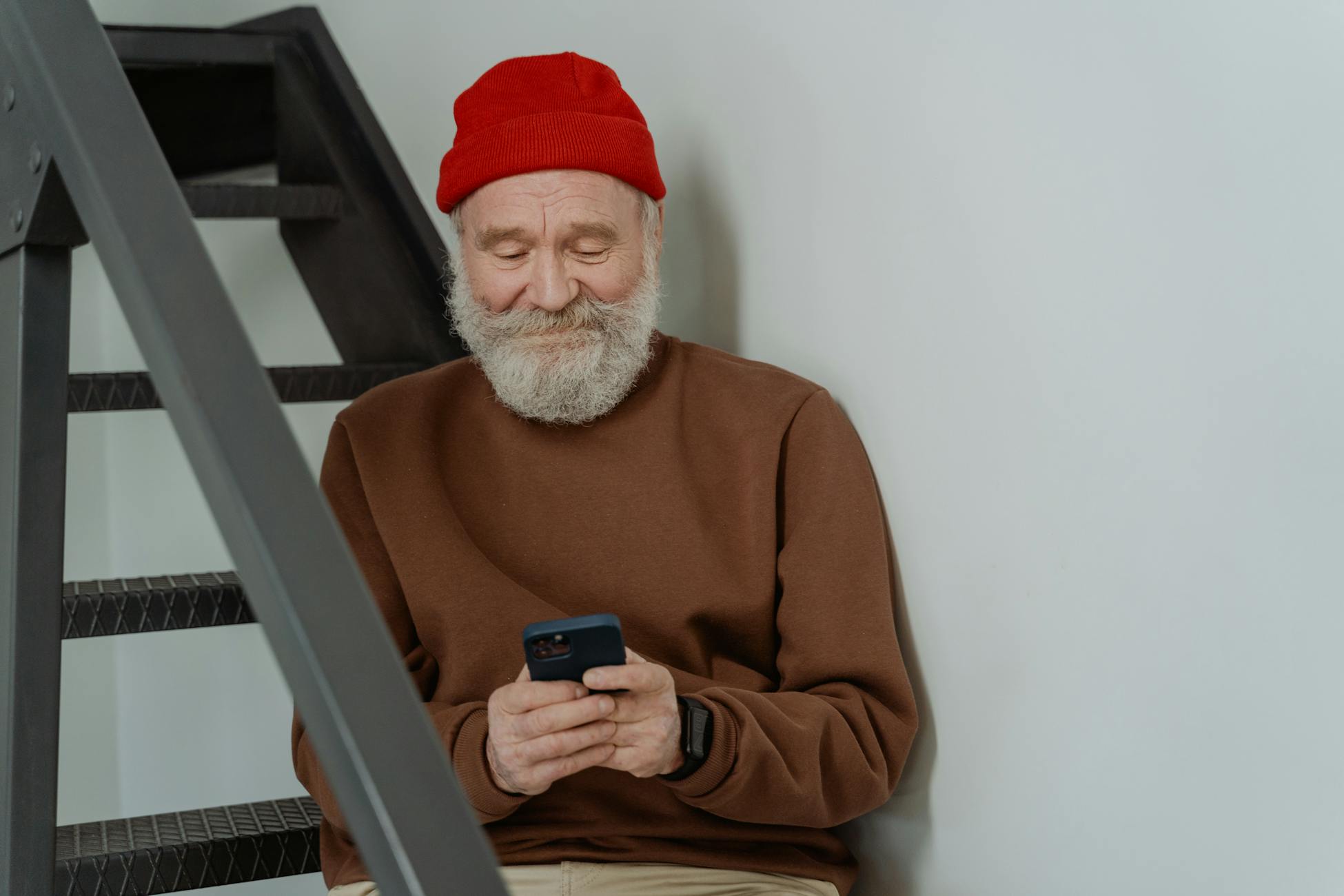 Senior man with beard using smartphone indoors, wearing a red beanie and brown sweater.