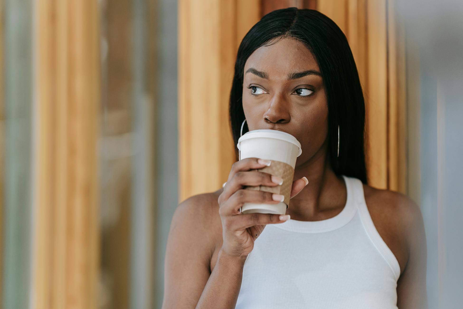 A woman in a white tank top thoughtfully sipping coffee indoors. Warm tones create a serene atmosphere.