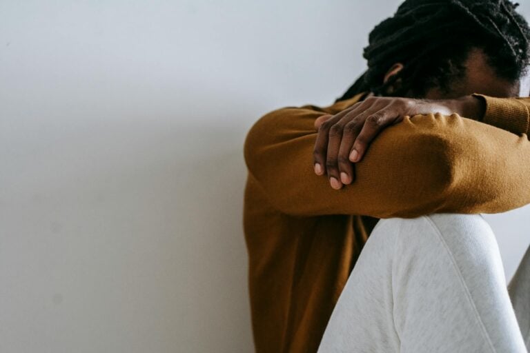 Crop anonymous depressed African American male embracing knees and covering face with arms while sitting on floor against white wall