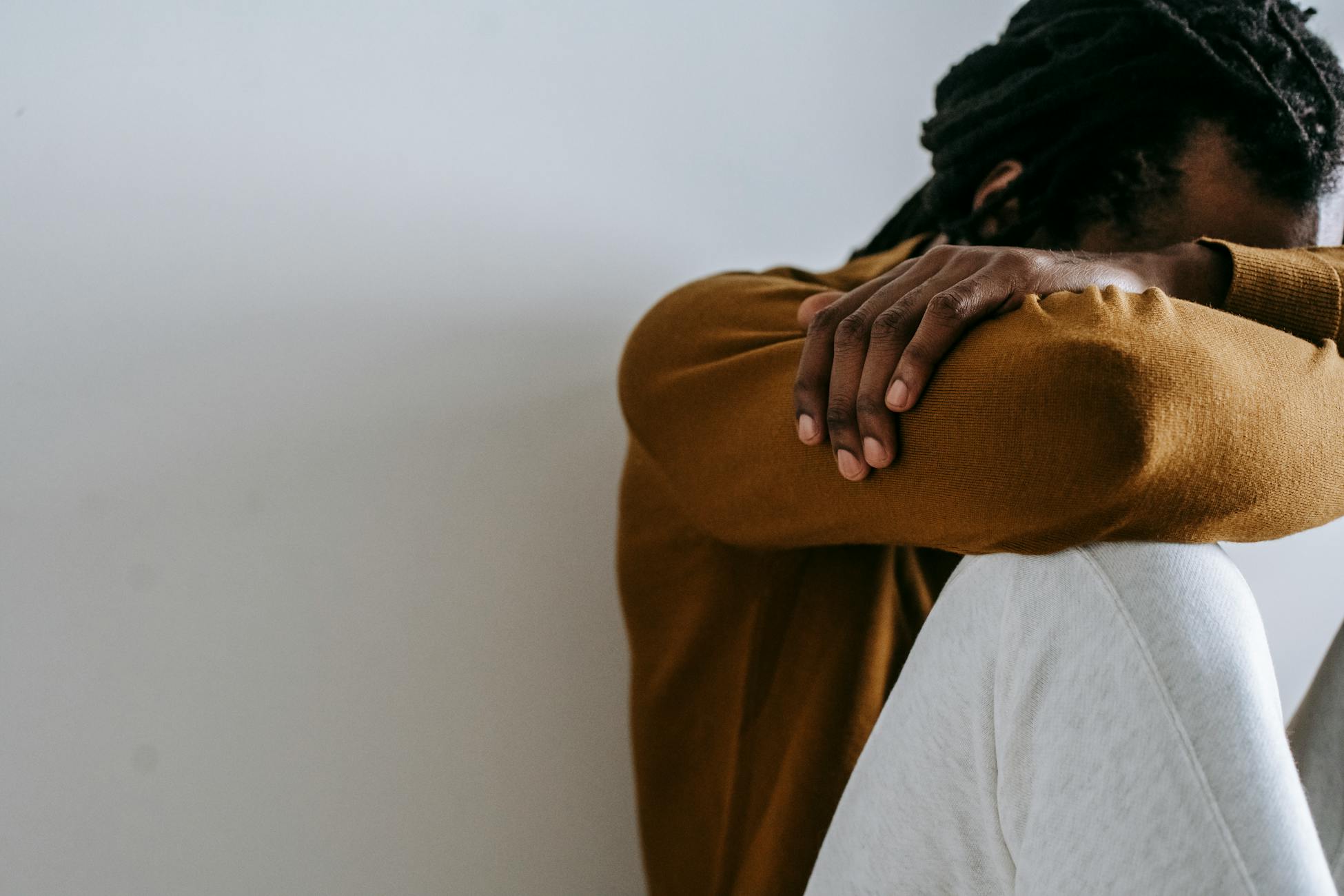 Crop anonymous depressed African American male embracing knees and covering face with arms while sitting on floor against white wall