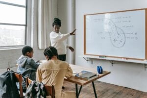 African American female teacher standing near whiteboard and giving lecture to ethnic teenagers during biology lesson