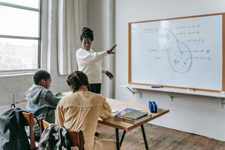African American female teacher standing near whiteboard and giving lecture to ethnic teenagers during biology lesson