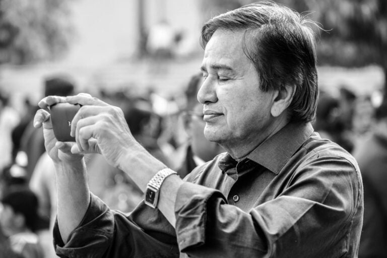 Elderly man in focus using smartphone camera outdoors, capturing memories in monochrome candid shot.