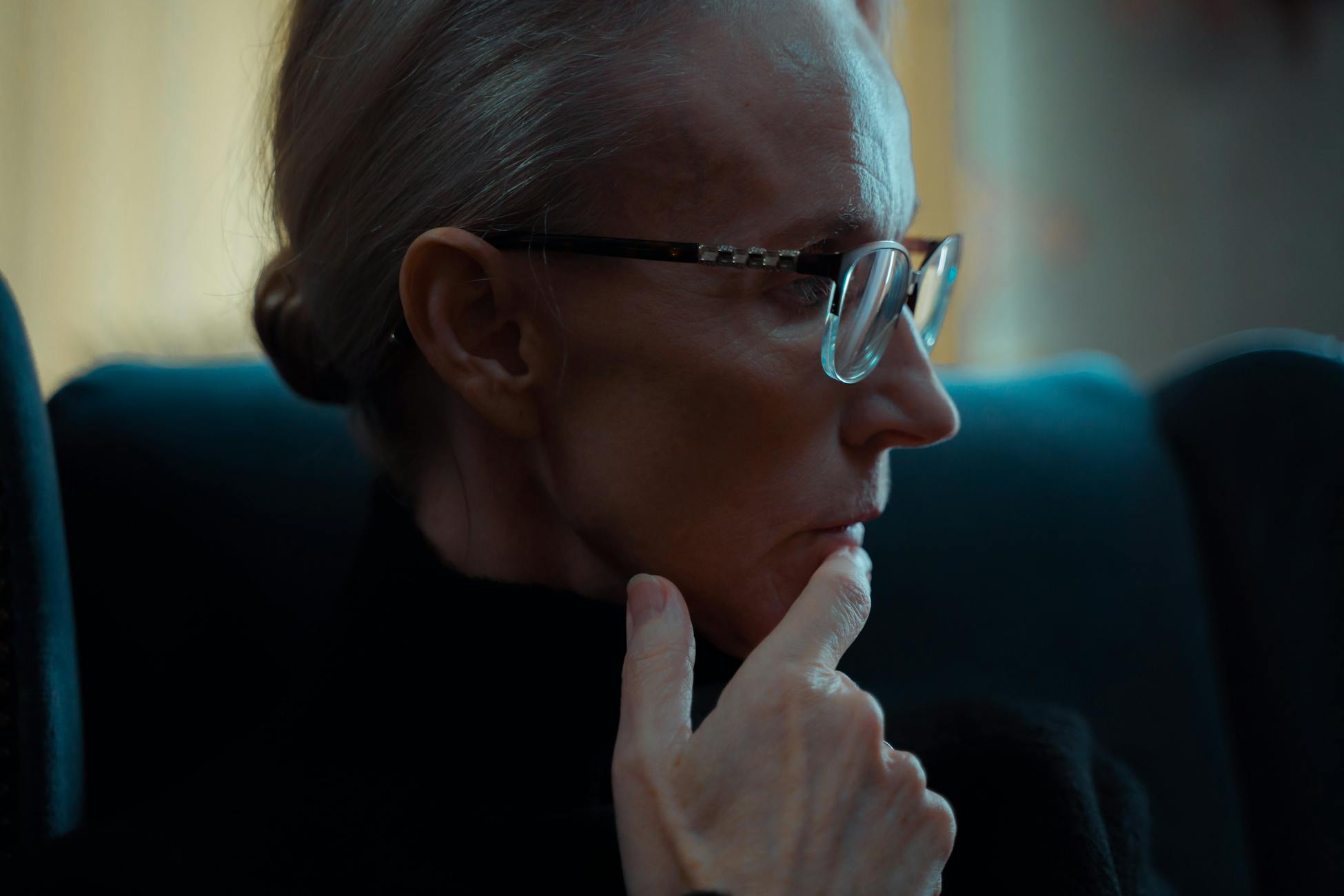 Side view of an elderly woman in glasses, deeply lost in thought indoors, with a muted ambiance.