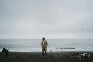 A solitary man walks on a cloudy beach with several dogs around him, conveying a moody and introspective atmosphere.