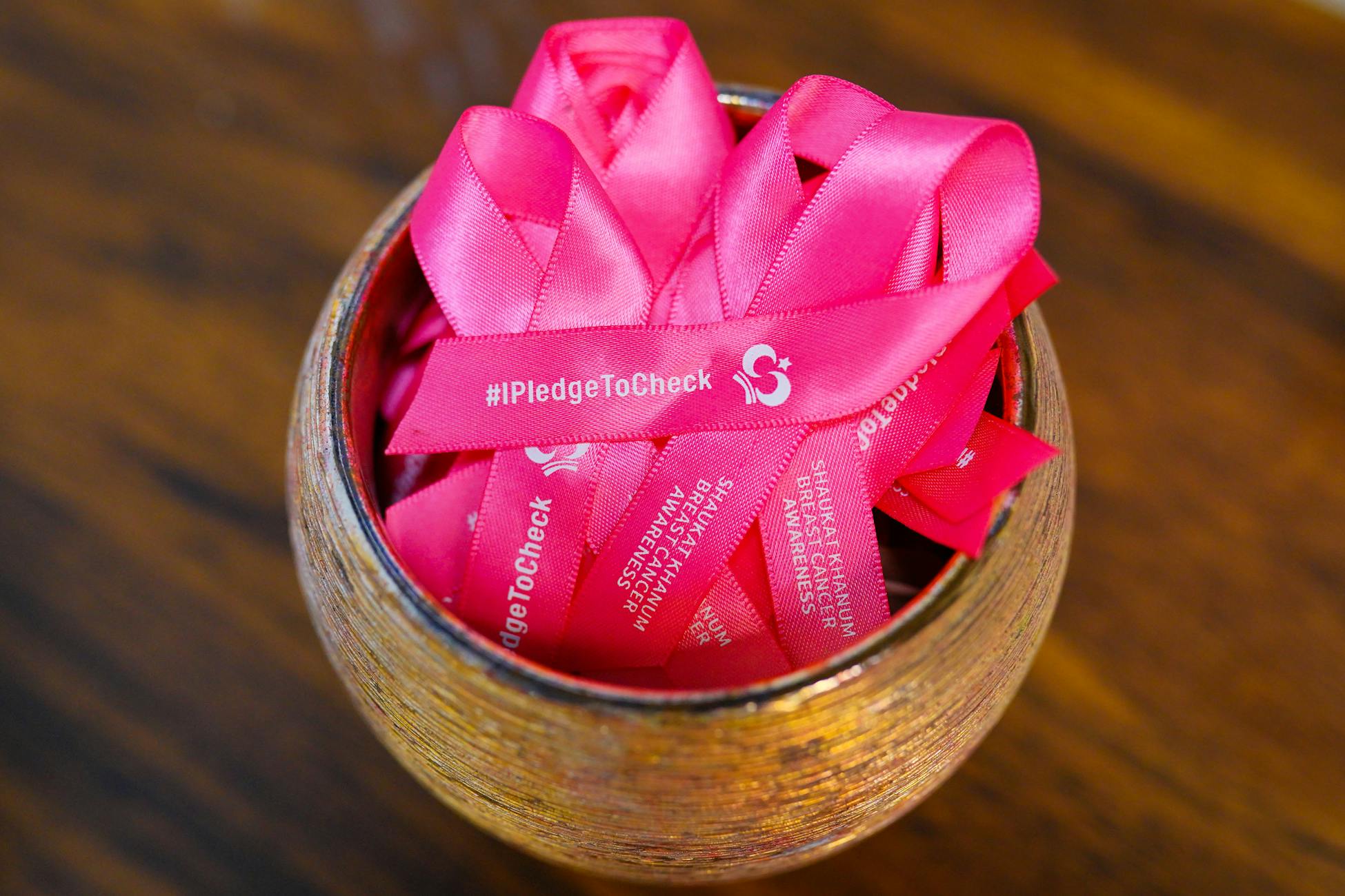 Pink ribbons for breast cancer awareness in a wooden bowl, promoting #IPledgeToCheck initiative.