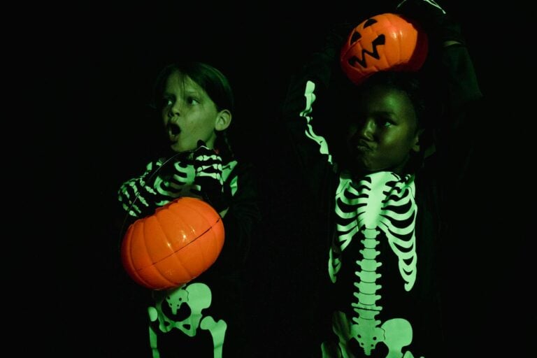 Two children in skeleton costumes holding jack-o'-lanterns, celebrating Halloween in a dark room.